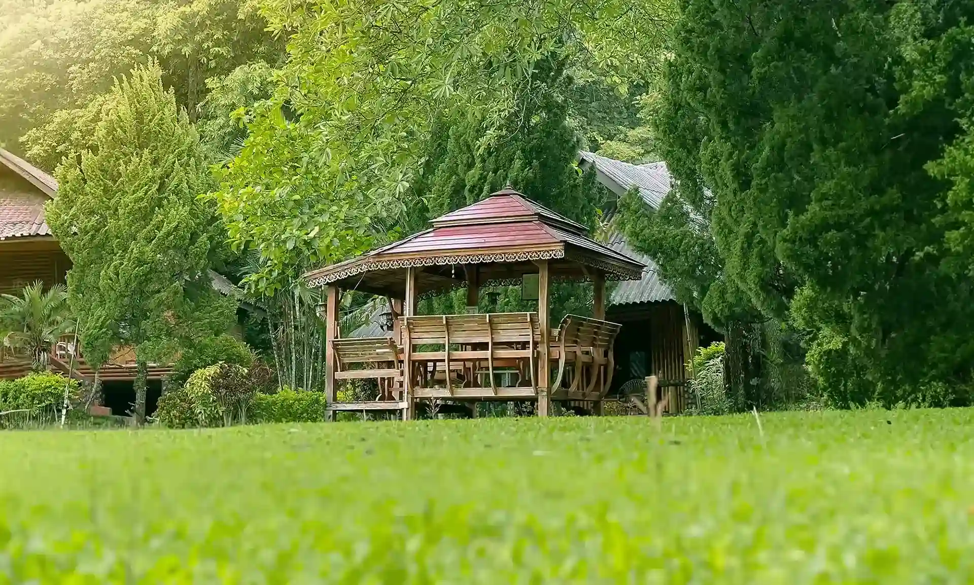 Open wooden pavilion in Siam Rehab garden with green lawn in foreground and sheer trees in background