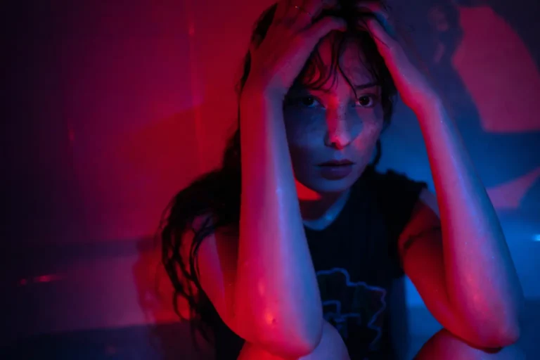 Woman seated with hands on her head under red and blue lighting, appearing distressed and overwhelmed.