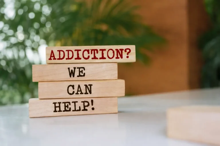 A stack of wooden blocks with the words "ADDICTION? WE CAN HELP!" stamped on them, set against a blurred background of greenery suggesting a therapeutic environment.