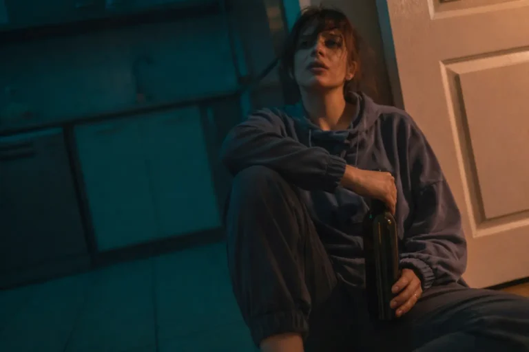 Woman seated on the floor holding a wine bottle, appearing withdrawn and contemplative in low lighting.