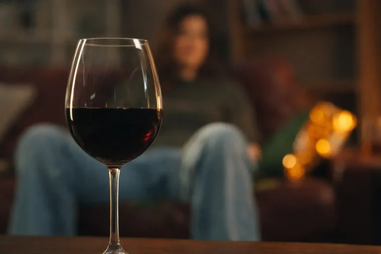 Glass of red wine in the foreground with a person seated in the background, illustrating alcohol presence within a domestic environment.