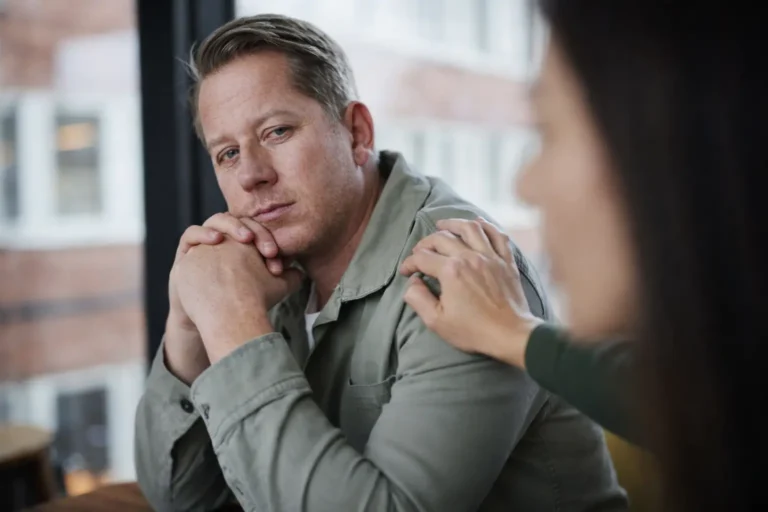 Man sitting at a table in a counseling-style setting while another person places a supportive hand on his shoulder during a recovery-related conversation.