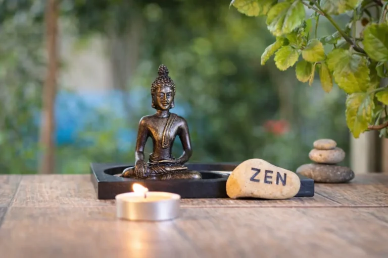 A bronze-style Buddha figurine sits on a dark tray next to a lit tea light candle and a stone with the word Zen printed on it.