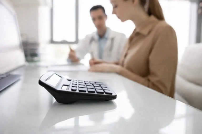 Close-up of a black calculator on a white desk, with a doctor and patient reviewing addiction treatment pricing documents in the blurred background.