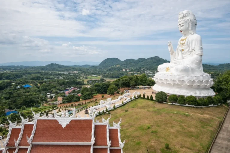 A massive white Guan Yin statue seated on a hilltop in Chiang Rai, Thailand, overlooking lush greenery and temple structures, illustrating the serene environment of international treatment destinations.