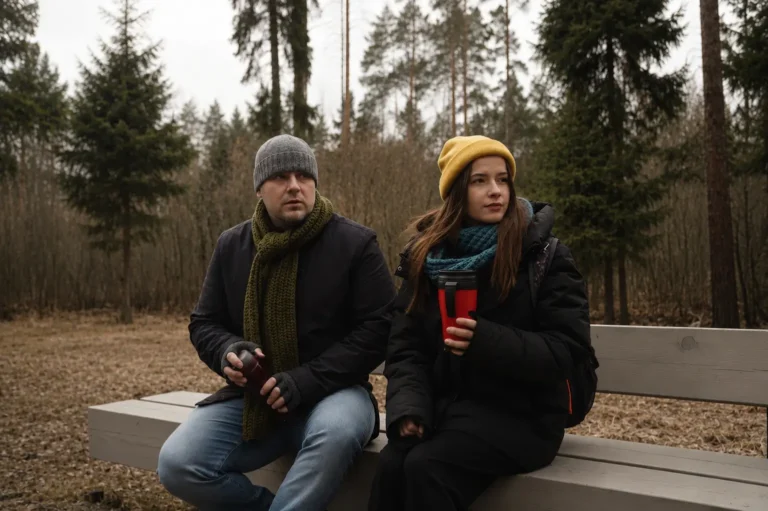 Mies ja nainen istuvat hiljaa penkillä talvisessa metsässä kahvikupit kädessä