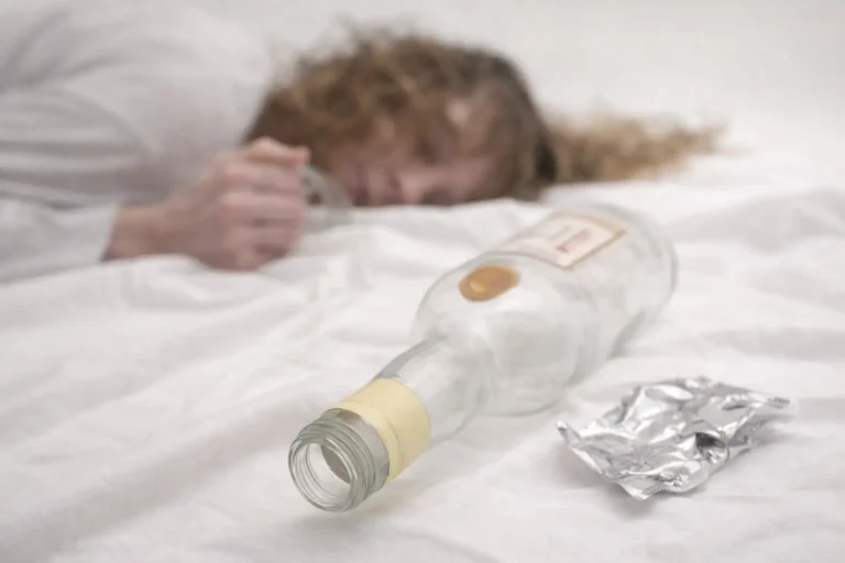 A person lies unconscious on white bedding with an empty liquor bottle in the foreground, illustrating a sign of relapse.