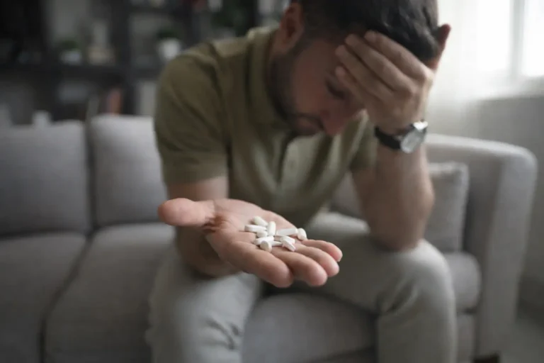 A distressed man sitting on a couch holding white pills in his hand, representing the despair and exhaustion of addiction relapse.