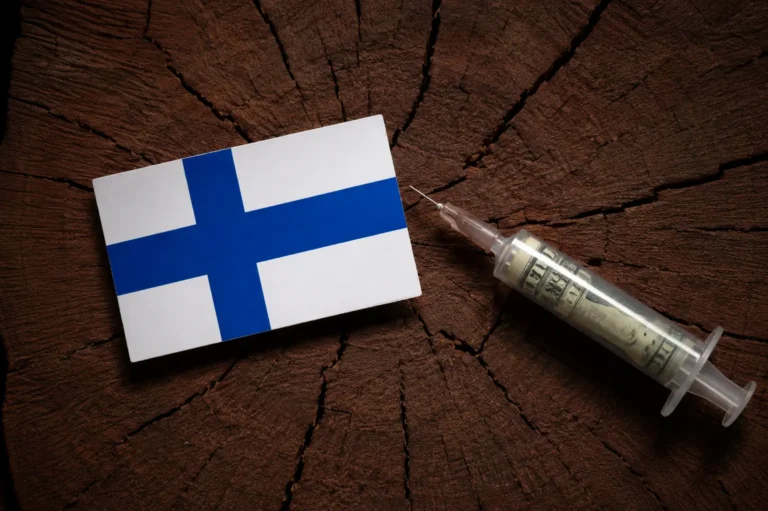 Suomen lippu ja rahatäytteinen ruisku puisen kannon pinnalla, kuvastamassa Suomen injektiohuumeongelmaa