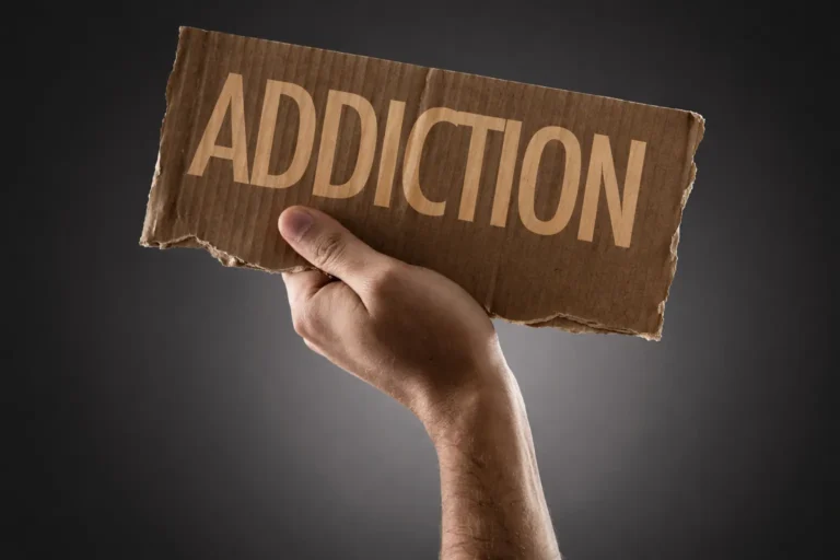 A person's hand holds up a torn piece of cardboard with the word "ADDICTION" printed on it against a dark background.