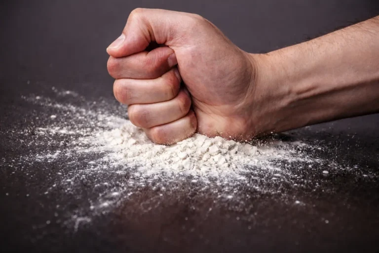Clenched fist striking a surface covered in white powder, dispersing dust particles, representing illicit substance use in a high-risk context.