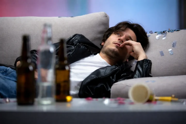 Man lying on a couch with alcohol bottles and pills on a table in front of him, appearing withdrawn and fatigued.
