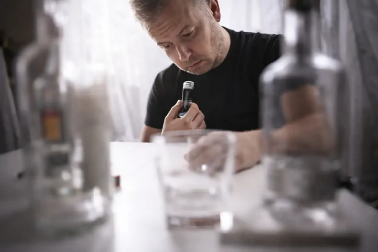 Middle-aged man intently examining a small bottle of alcohol at a table with empty glasses, illustrating isolation and contemplation often associated with alcohol dependence in a private home setting.