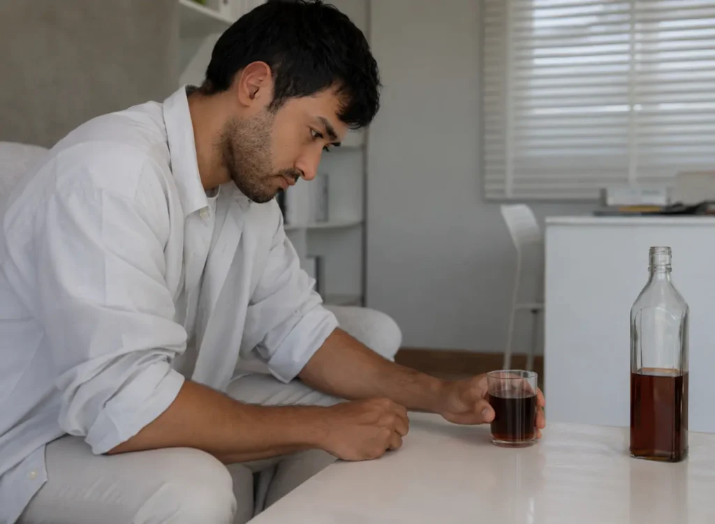 Man sitting alone at a kitchen table holding a glass of whiskey, looking down - struggling with alcohol addiction