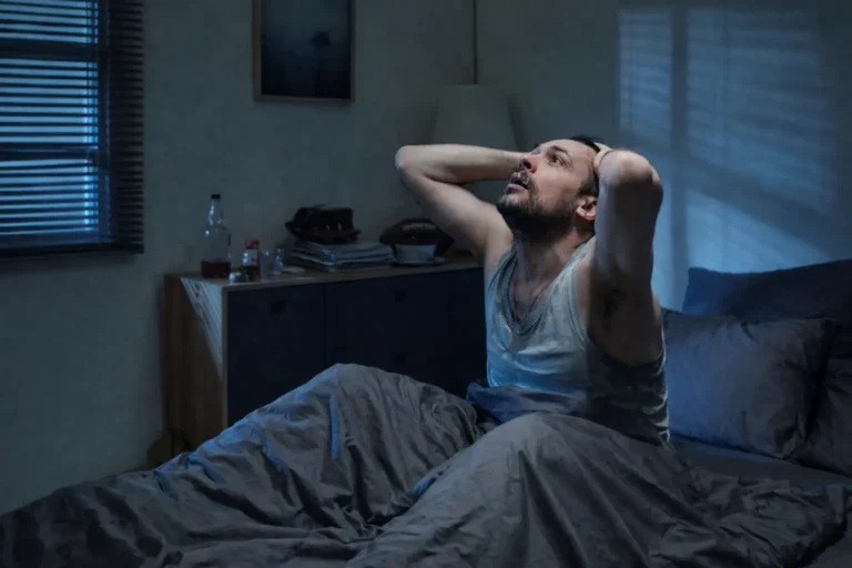 A man in distress sits up in bed at night, with a bottle of alcohol on the nightstand, symbolizing the pain of a relapse after detox.