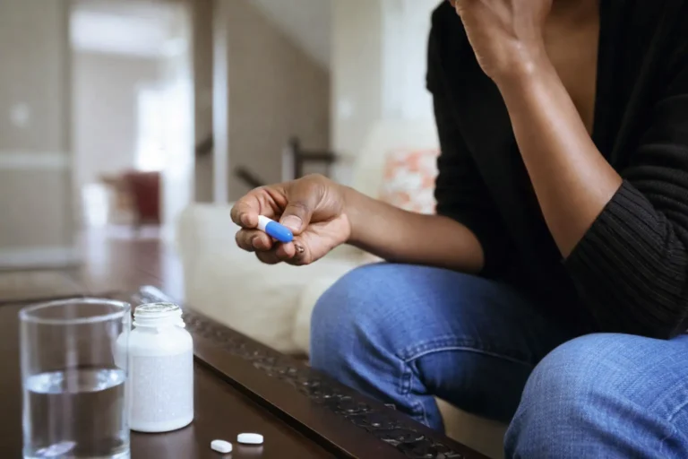 Close-up of a person holding a blue and white capsule near a water glass and pill bottle, illustrating medication management boundaries in early recovery.
