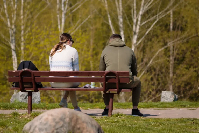 A mother and adult son sitting apart on a park bench in tense silence, illustrating the emotional distance when a loved one refuses rehab.
