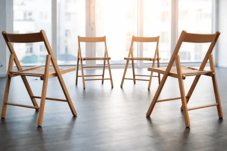 Four wooden chairs arranged in a circle in a bright outpatient group therapy room with large windows.