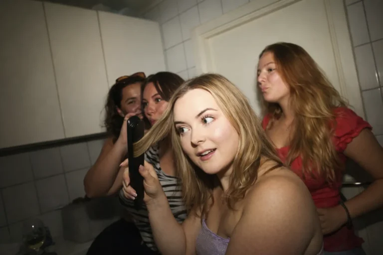 Group of young women gathered in a bathroom during a social event, preparing together in a close, high-energy setting.