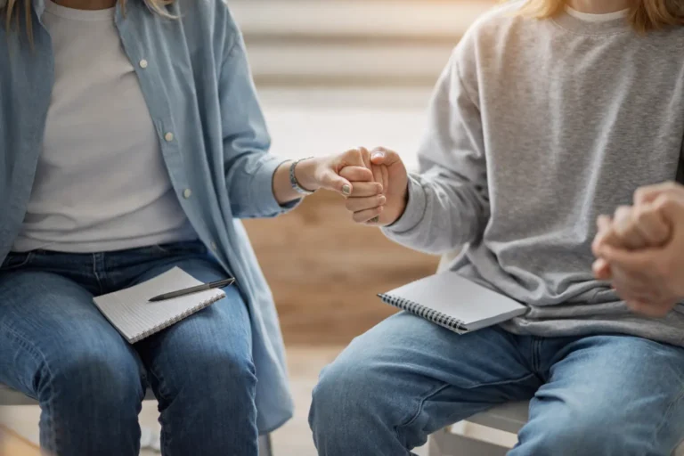 Two individuals seated side by side holding hands during a group support session, representing structured rehabilitation care within Australia's private and public treatment systems.