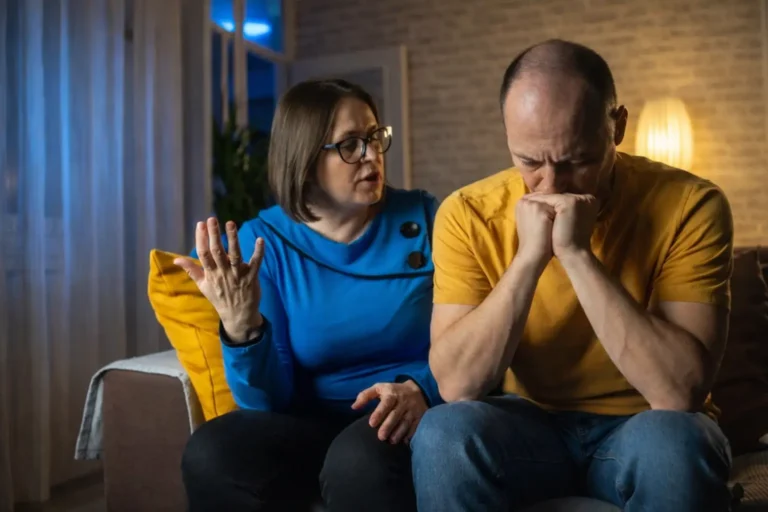 A woman talks to a man who is sitting with his head in his hands, depicting a high-stress conversation about seeking addiction treatment while balancing career concerns.