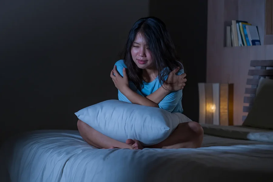 Distressed woman sitting on bed at night, hugging herself, showing signs of psychotic disorder struggles.