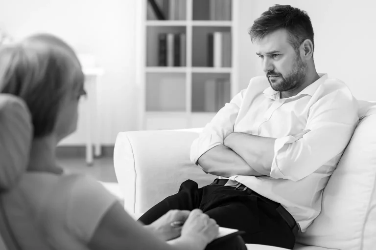 A man sits with folded arms on a couch during a counseling session, reflecting on struggles with addiction, while a therapist listens attentively at Siam Rehab.