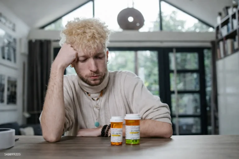 A tired young man sitting at a table staring at two prescription pill bottles, illustrating the exhaustion and decision fatigue common in early recovery stabilization.