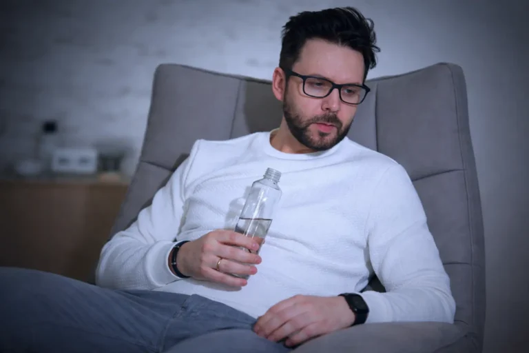 Man sitting in a chair holding a bottle and looking down, reflecting relapse patterns, withdrawal from supports, and post-treatment vulnerability requiring risk reassessment and boundary recalibration.