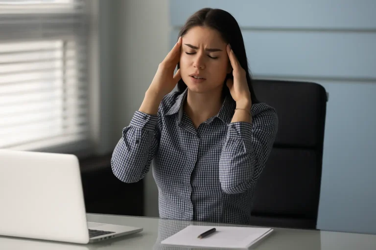 Woman sitting at a desk with a laptop, holding her head in stress and discomfort, symbolizing the struggles of anxiety, burnout, or substance dependence and the need for professional recovery support at Siam Rehab.