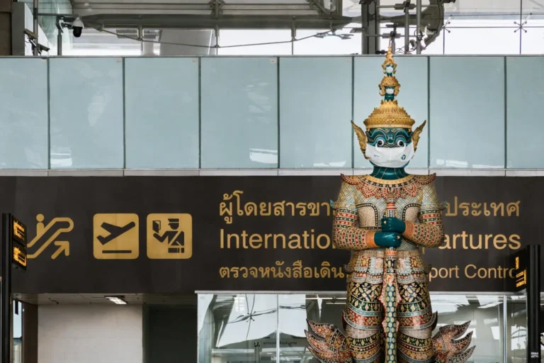 A large traditional Thai Yaksha guardian statue wearing a white protective mask stands in front of the International Departures and Passport Control signage at an airport.