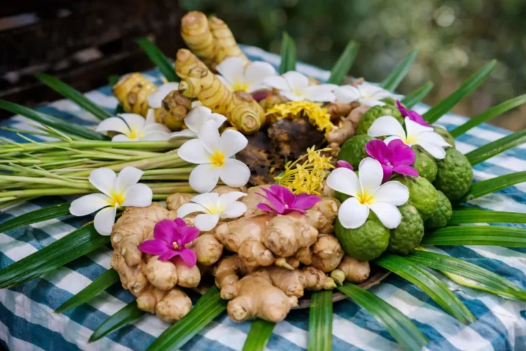 Fresh ginger root, kaffir limes, lemongrass stalks, and white frangipani flowers arranged on a blue checkered tablecloth.