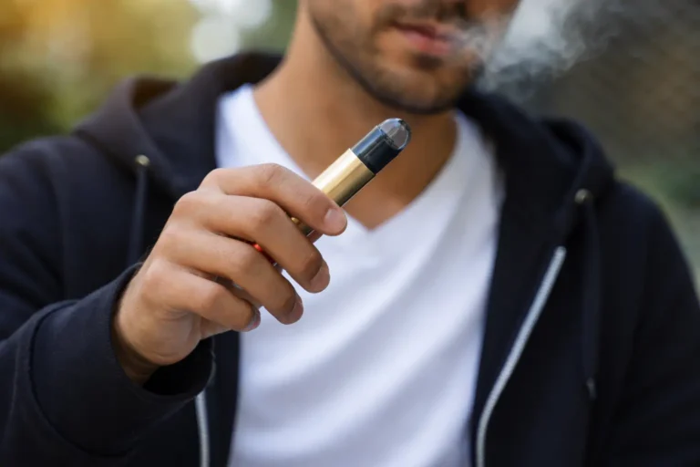 Man holding and using a vape device outdoors with visible vapor, representing nicotine or substance use behavior.