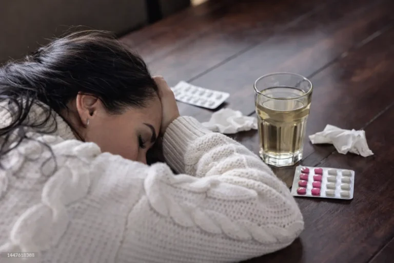 Woman slumped over table with a blister pack of pills and a glass of alcohol, illustrating the physical danger and emotional exhaustion of chronic relapse.