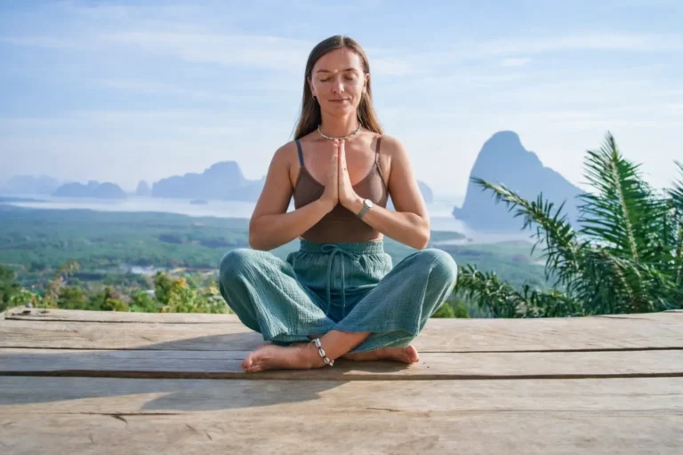 A woman sits cross-legged with hands together on a wooden platform overlooking a tropical bay with mountains.
