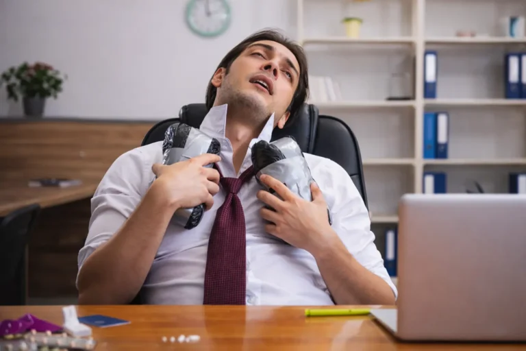 Office worker seated at a desk holding cold packs to his neck, with pills visible on the table, illustrating stress-related substance use risk in a professional setting.
